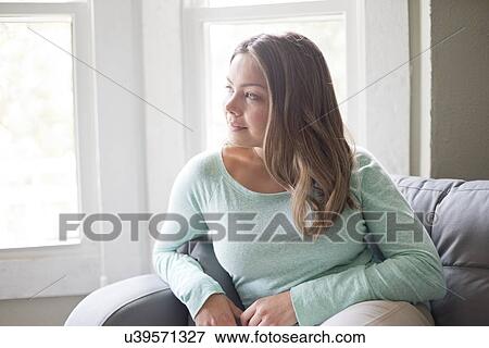 Stock Photo - Young woman sitting on sofa and looking away.. Fotosearch