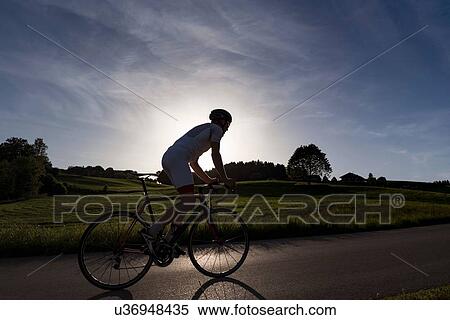 Stock Photography - Backlit view of young male cyclist cycling up rural road at sunset. Fotosearch