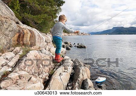 Stock Image - Boy standing on rock by fjord playing with toy boat, Aure, More og Romsdal, Norway. Fotosearch