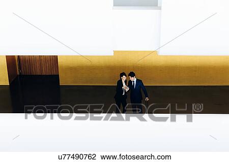 Stock Image - Businessman and businesswoman with wheeled luggage in hotel building. Fotosearch