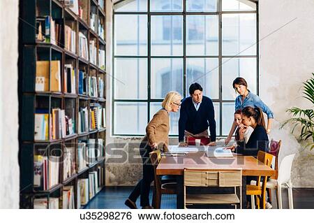 Stock Photograph - Businessman and businesswomen gathered around table, looking at books. Fotosearch