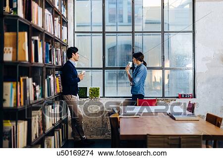 Picture - Businessman and woman standing beside window, drinking from disposable coffee cups, in conversation. Fotosearch