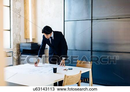 Stock Image - Businessman in office, making notes on blueprints. Fotosearch