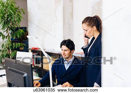 Stock Photography - Businessman sitting at desk, woman standing beside him using smartphone. Fotosearch