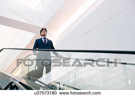 Stock Image - Businessman with wheeled luggage on hotel escalator. Fotosearch