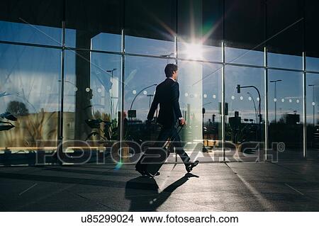 Picture - Businessman with wheeled luggage passing glass building, Malpensa, Milan. Fotosearch