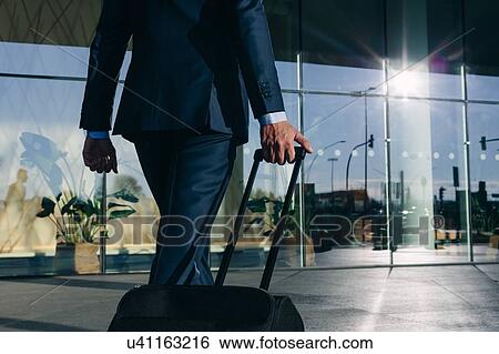 Stock Photograph - Businessman with wheeled luggage walking towards glass building, Malpensa, Milan. Fotosearch