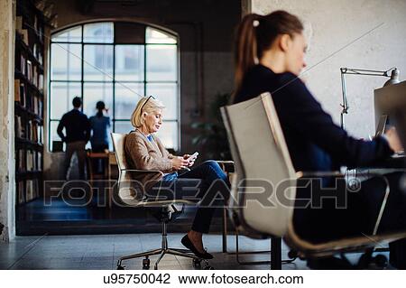 Stock Image - Businesspeople in office, two women sitting at desk working, businessman and woman looking out of window in background. Fotosearch