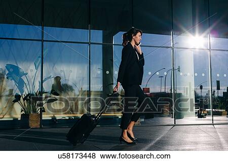 Stock Photo - Businesswoman with wheeled luggage passing glass building, Malpensa, Milan. Fotosearch