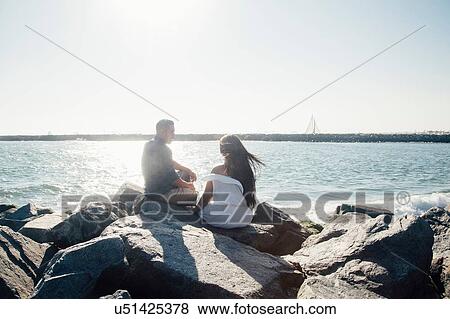 Couple sitting on coastal rocks, looking at view, rear view View Large Photo Image Stock Photo - Couple sitting on coastal rocks, looking at view, rear view. Fotosearch