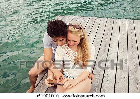 Couple sitting on pier looking at smartphone View Large Photo Image Stock Image - Couple sitting on pier looking at smartphone. Fotosearch
