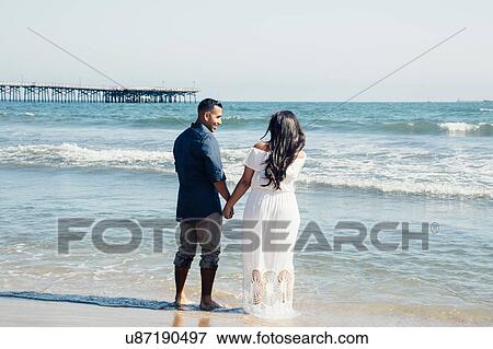 Stock Photo - Couple standing on beach, holding hands, rear view, Seal Beach, California, USA. Fotosearch