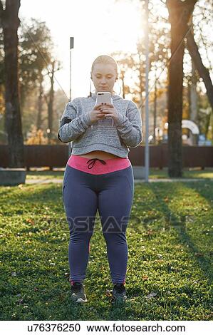 Stock Image - Curvaceous young woman training in park, looking at smartphone. Fotosearch