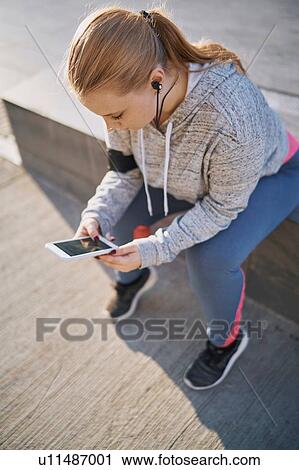 Stock Image - Curvaceous young woman training, sitting on wall looking at smartphone. Fotosearch