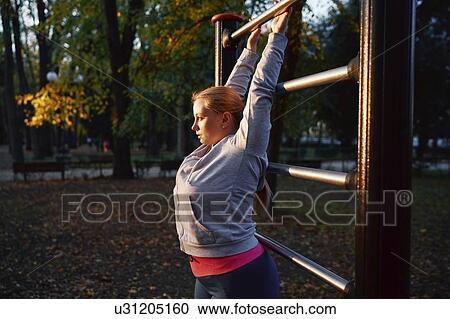 Stock Image - Curvaceous young woman training, gripping exercise bar in park. Fotosearch