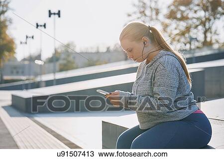 Stock Image - Curvaceous young woman training, sitting on wall looking at smartphone. Fotosearch