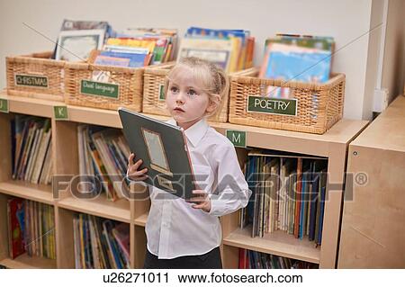 Stock Image - Cute schoolgirl holding book in classroom at primary school. Fotosearch