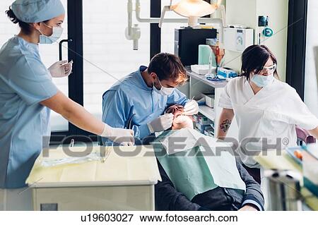 Dentist looking into male patient's mouth, dental nurses preparing equipment View Large Photo Image Stock Photo - Dentist looking into male patient's mouth, dental nurses preparing equipment. Fotosearch
