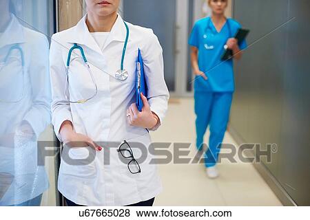 Doctor standing in hospital hallway, surgeon walking in background, mid section View Large Photo Image Stock Photo - Doctor standing in hospital hallway, surgeon walking in background, mid section. Fotosearch