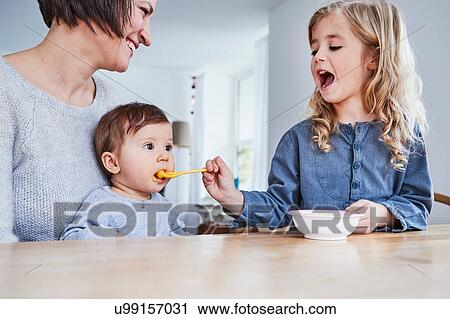 Family sitting at kitchen table, young girl spoon-feeding baby sister View Large Photo Image Stock Image - Family sitting at kitchen table, young girl spoon-feeding baby sister. Fotosearch