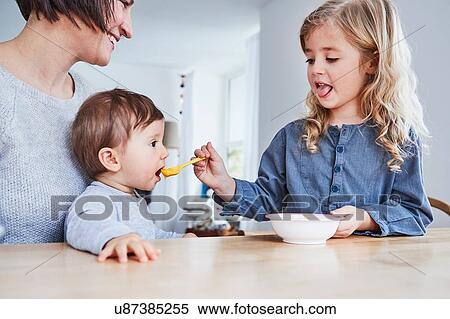 Stock Photography - Family sitting at kitchen table, young girl spoon-feeding baby sister. Fotosearch