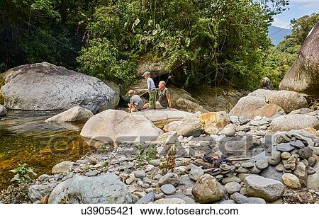Stock Image - Father and sons relaxing on rocks beside water, Ventilla, La Paz, Bolivia, South America. Fotosearch