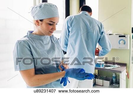 Stock Photo - Female dentist in dentist office, putting on surgical gloves, male dentist in background. Fotosearch