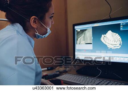Stock Image - Female dentist looking at model of teeth on computer screen. Fotosearch