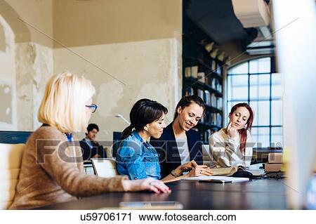 Four businesswomen sitting at desk in office, looking through book View Large Photo Image Stock Image - Four businesswomen sitting at desk in office, looking through book. Fotosearch