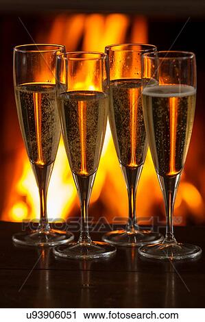 Stock Image - Four filled champagne glasses on table in front of burning fire. Fotosearch