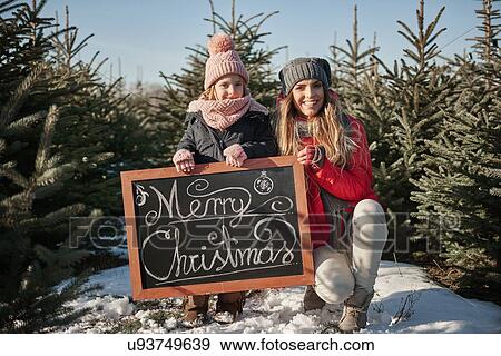 Stock Photo - Girl and mother in christmas tree forest with merry christmas sign, portrait. Fotosearch