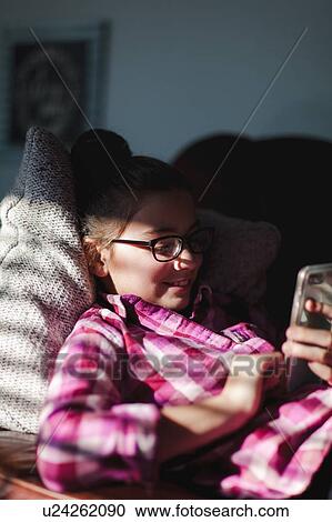 Stock Image - Girl relaxing on sofa, looking at smartphone. Fotosearch