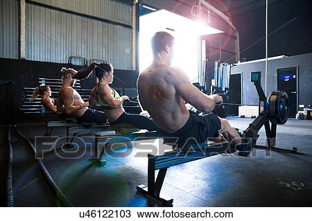 Stock Image - Group of people exercising in gymnasium, using rowing machines, rear view. Fotosearch