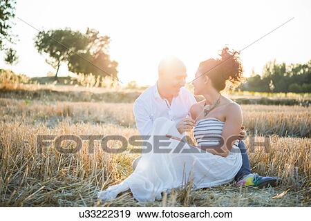 Stock Photo - Heterosexual couple sitting in field, face to face, smiling. Fotosearch