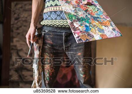 Stock Photo - Male artist holding palette and paint brush in artists studio, mid section. Fotosearch