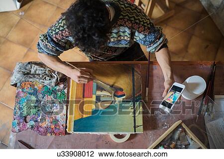 Stock Image - Male artist looking at smartphone while painting canvas in artists studio, overhead view. Fotosearch