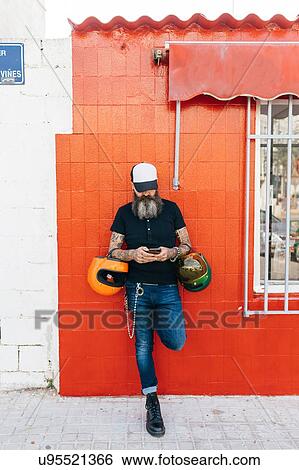Male hipster leaning against red wall looking at smartphone View Large Photo Image Stock Photograph - Male hipster leaning against red wall looking at smartphone. Fotosearch