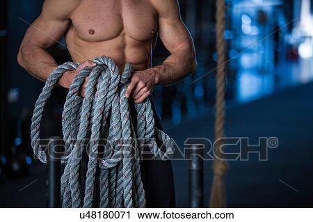 Stock Image - Man exercising in gymnasium, holding rope, preparing for sled training, mid section. Fotosearch