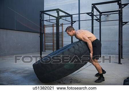 Stock Image - Man exercising in gymnasium, lifting tire. Fotosearch