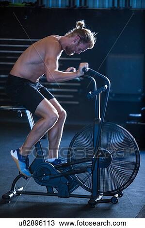 Stock Image - Man exercising in gymnasium, using air resistance exercise bike. Fotosearch