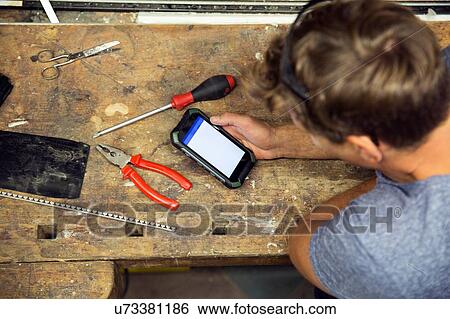 Stock Photograph - Man in workshop, using smartphone, overhead view. Fotosearch