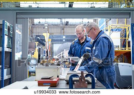 Stock Image - Mature and younger engineers inspecting turbine blade in turbine maintenance factory. Fotosearch