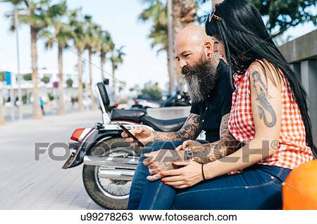 Stock Image - Mature hipster couple on bench looking at smartphone, Valencia, Spain. Fotosearch