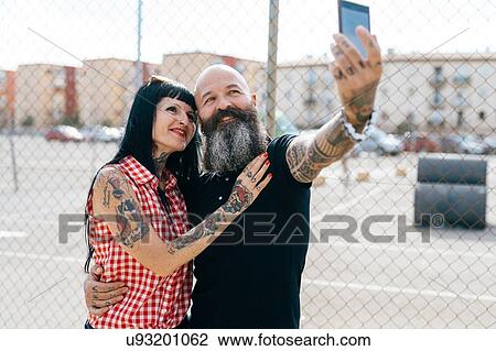 Stock Image - Mature hipster couple taking selfie by wire fence. Fotosearch