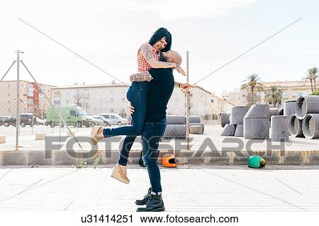 Stock Image - Mature hipster man lifting and hugging girlfriend in city. Fotosearch