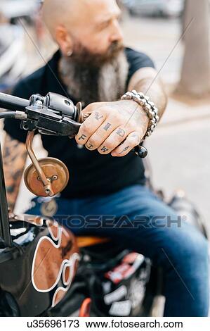 Stock Image - Mature male hipster astride motorcycle, close up of tattooed hand. Fotosearch