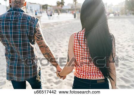 Stock Image - Mature tattooed hipster couple strolling on beach, rear view, Valencia, Spain. Fotosearch
