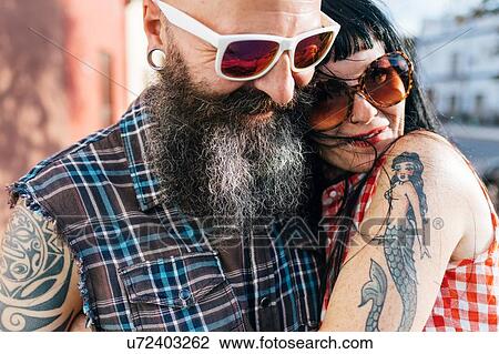 Stock Image - Mature tattooed hipster couple embracing on sidewalk, close up. Fotosearch