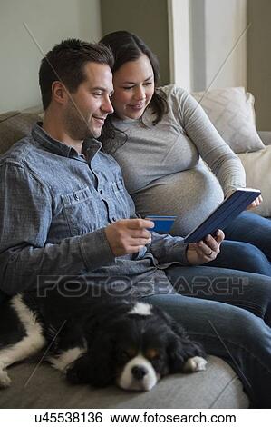 Stock Photograph - Mid adult couple shopping online in living room, sitting on sofa with puppy. Fotosearch