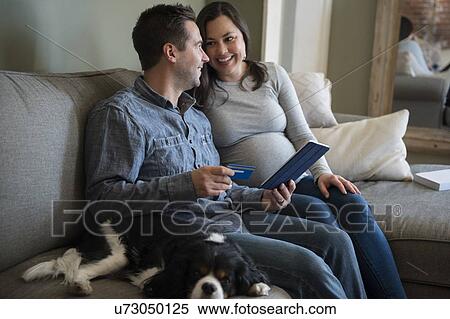 Stock Photography - Mid adult couple shopping online in living room, sitting on sofa with puppy. Fotosearch